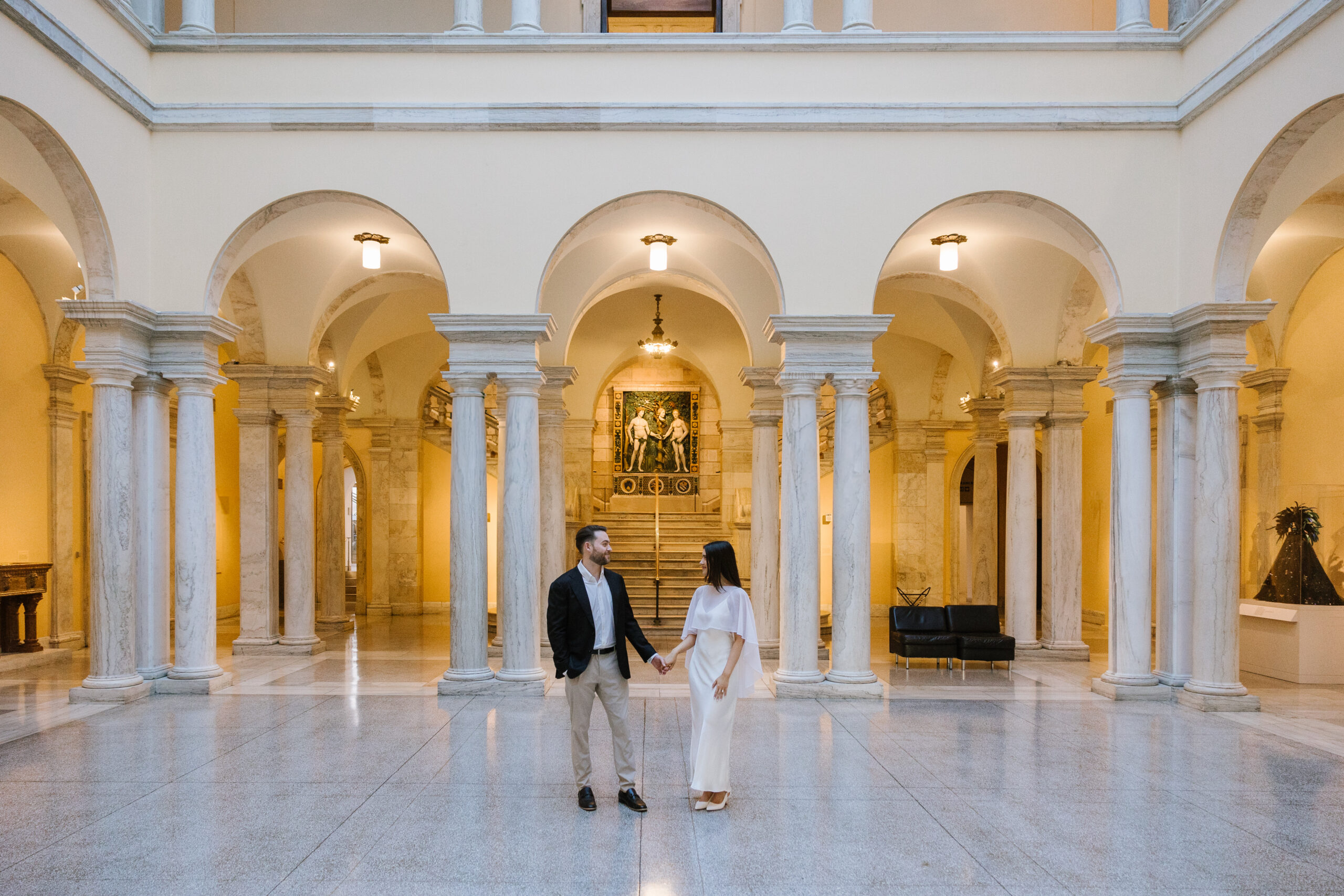 engaged couple stands holding hands in middle of marble columned atrium at walters art museum