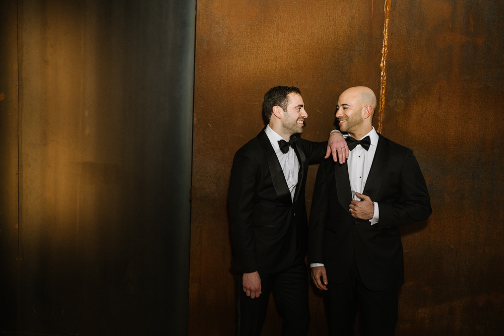 just married husbands standing next to each other in black tuxes against metal wall at the butterfly room in baltimore